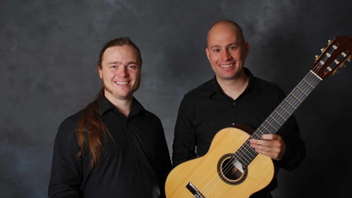 To mænd står sammen, hvor den ene holder en guitar.