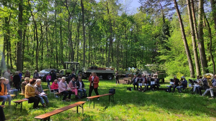 Auf einer Waldlichtung sitzen Menschen auf Bänken. Etwas abseits sitzt der Posaunenchor. Im Hintergrund Traktoren mit Anhängern zum Sitzen.
