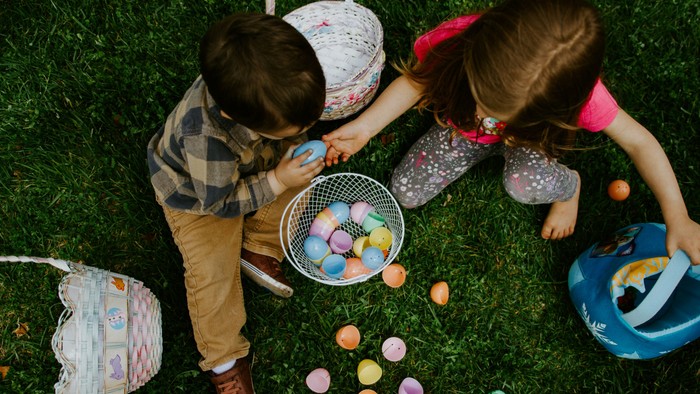Zwei Kinder sitzen auf Gras und sammeln bunte Ostereier in Körben.