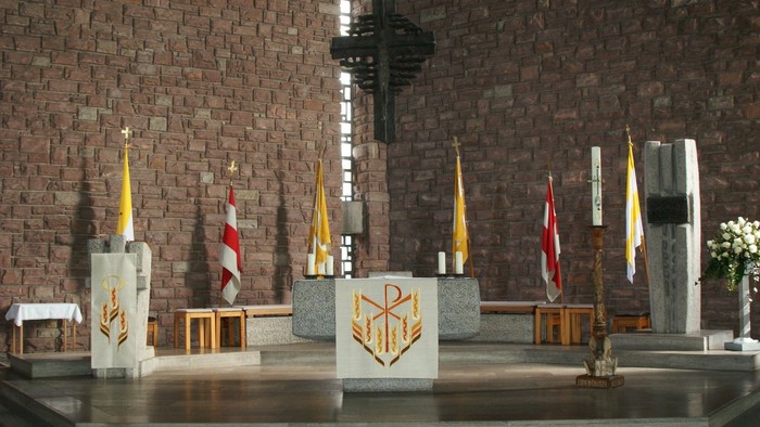 Religiöser Altarbereich mit Flaggen, Kerzen und einem markanten Kreuz vor einem Ziegelhintergrund.
