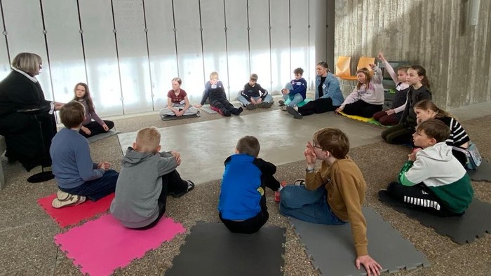 Børn sidder i kreds på yogaunderlag i et rum med store vinduer