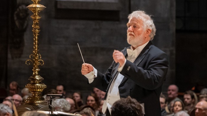 En dirigent leder et orkester, mens han holder en taktstok, med en gylden lysestage i nærheden.