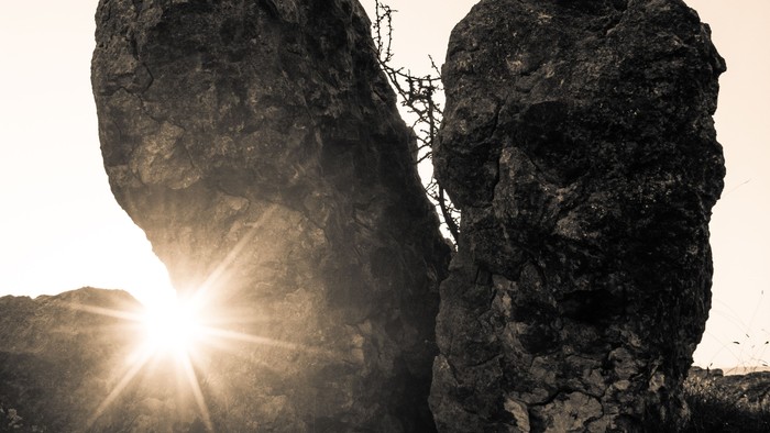 To majestætiske klippeformationer tegner sig som silhuetter mod en glødende solnedgangshimmel med solstråler, der bryder frem.