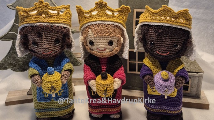 Tre hæklede figurer med kroner, der hver holder en gave, placeret foran et træhus og en juletræ.