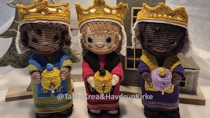 Tre hæklede figurer med kroner, der hver holder en gave, placeret foran et træhus og en juletræ.