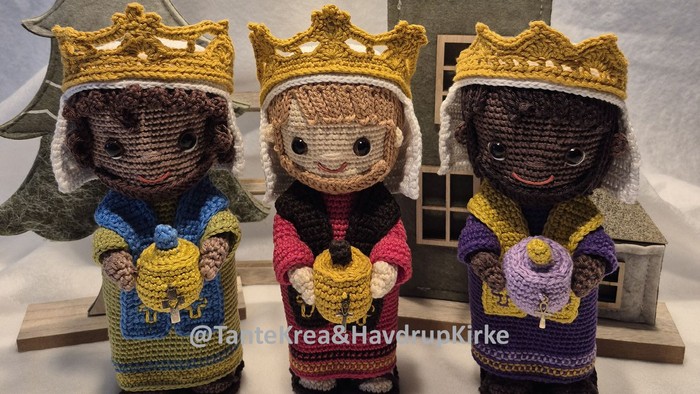 Tre hæklede figurer med kroner, der hver holder en gave, placeret foran et træhus og en juletræ.