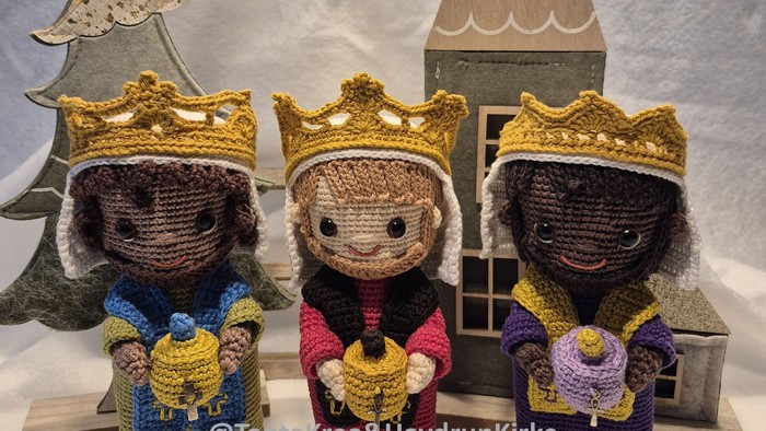 Tre hæklede figurer med kroner, der hver holder en gave, placeret foran et træhus og en juletræ.
