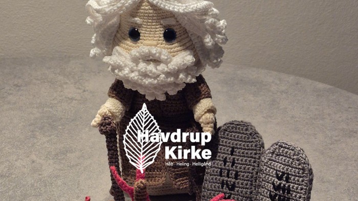 Hæklet figur, der holder et farverigt kors med gravsten og hat, som repræsenterer et religiøst tema.