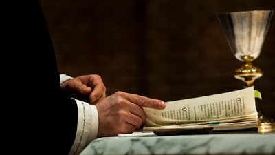 En person holder et håndskrift og sitter ved et alter med en kelch.