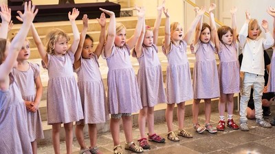 En gruppe børn i lilla tøj står med hænderne op i en kirke.