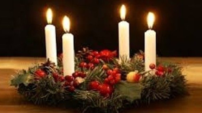 En adventskranse med fire tændte lys og pynt med grønne nåle og rodgrønne bær.