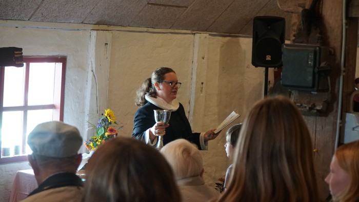 En taler henvender sig til et opmærksomt publikum i en rustik indendørs ramme med papirer og en mikrofon.