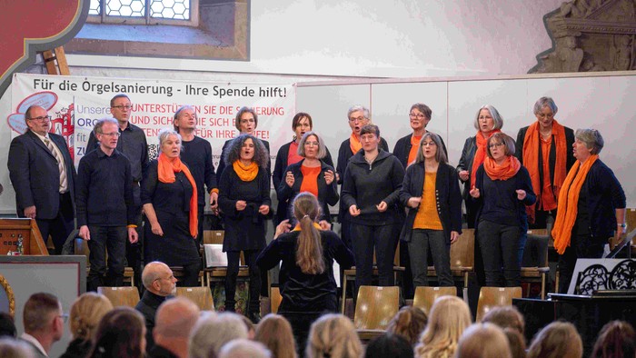 Ein Chor tritt in einer Kirche auf und sammelt Spenden für die Restaurierung der Orgel, unterstützt vom Publikum und einem Spendenaufruf-Banner.