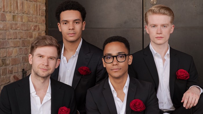 Vier Männer in formeller Kleidung mit roten Boutonnières posieren gemeinsam drinnen.