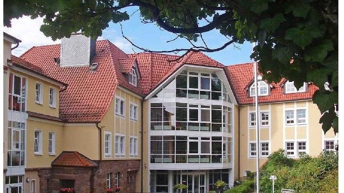 Das Bild zeigt ein großes, gelbes Gebäude mit rotem Ziegeldach und großen Fenstern, umgeben von Bäumen.