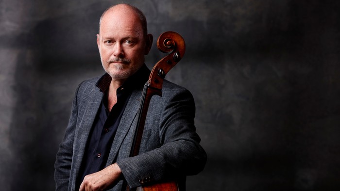 En skaldet mand i jakkesæt poser med en cello hvilende på sin skulder.