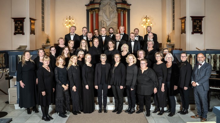 En formel kor- og orkestergruppe poserende sammen i et stort kirkeinteriør.
