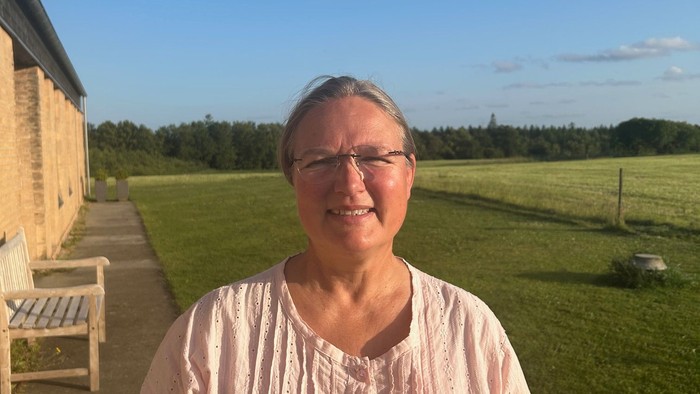 En kvinde står i en have med en mur og skov i baggrunden