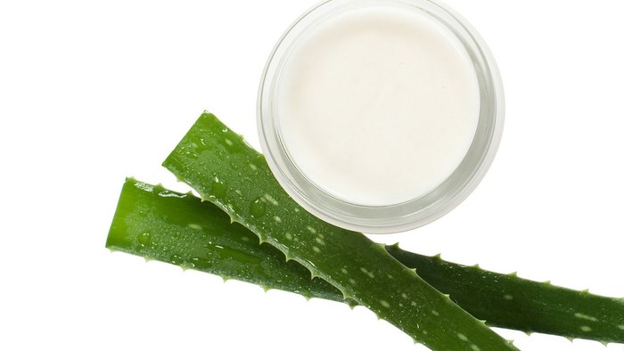Ein mit weißer Creme gefüllter Glasbehälter steht neben zwei frischen, grünen Aloe-Vera-Blättern auf weißem Hintergrund.