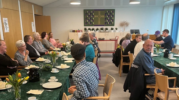 En gruppe mennesker samles til et fælles måltid ved pyntede borde med blomsterdekorationer i en lys spisesal.