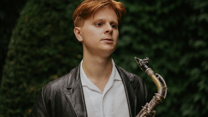 En ung mand med rødt hår holder en saxofon udendørs.