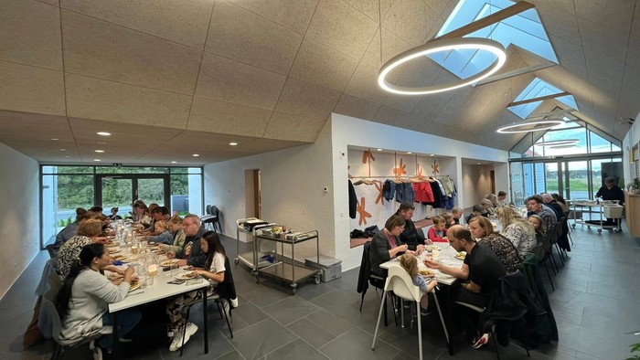 Mennesker, der spiser sammen i et moderne, rummeligt indendørs arrangementrum med store vinduer og stilfuld belysning.