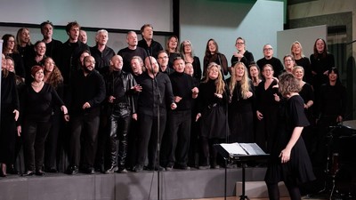 En stor korgruppe på scenen, alle i sort tøj, med en dirigent foran.