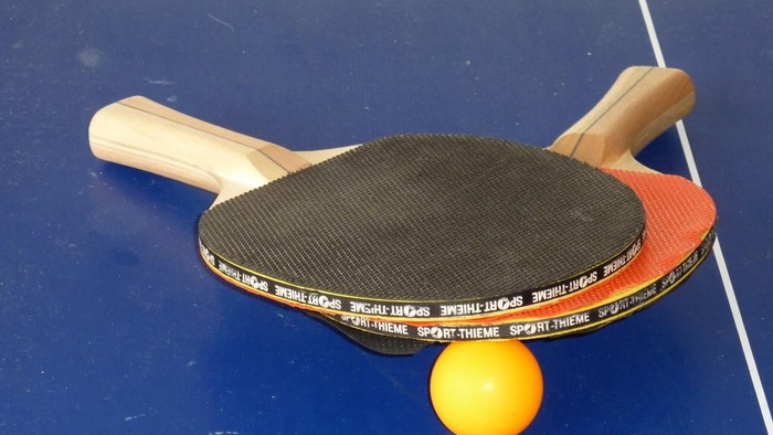 Tischtennisschläger mit schwarzer Gummioberfläche und ein oranger Ball auf einer blauen Tischfläche.