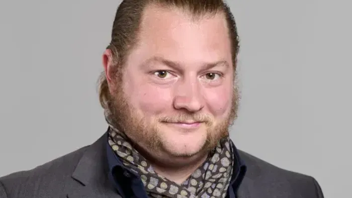 Mann mit Bart und Schnurrbart, der einen Anzug und ein gemustertes Halstuch trägt.