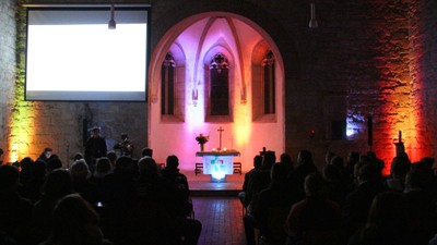 dunkler Kirchenraum, in buntes Licht getaucht, Altar mit Kerzen, Kreuz und Blumen, Personen von hinten sitzend, Leinwand 
