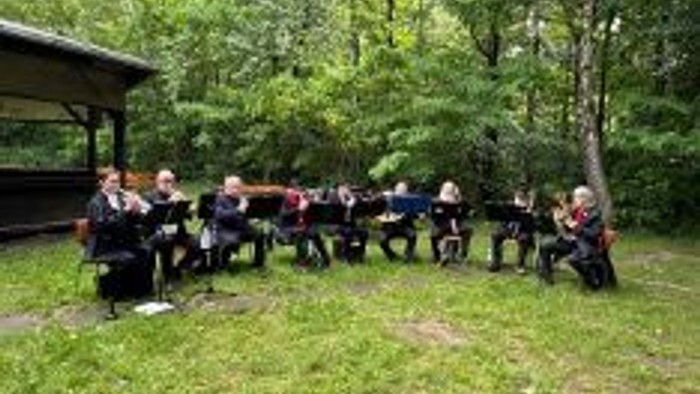 Gruppe von Menschen, die im Freien auf einer Rasenfläche in der Nähe eines Pavillons sitzen und gemeinsam Musikinstrumente spielen.