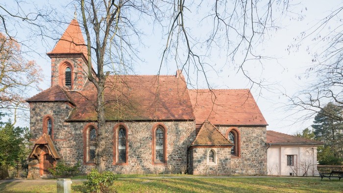Ein steinernes Kirchgebäude mit rotem Dach und Turm, umgeben von Bäumen und Gras.