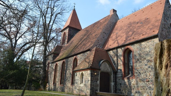 Kleine Kirche mit rotem Dach und Steinmauern, daneben eine Bank auf dem Rasen