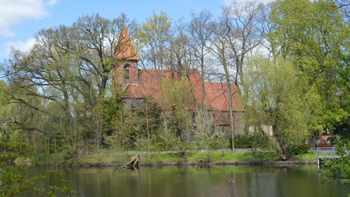 Ein rotes Backsteingebäude mit spitzem Turm steht hinter Bäumen und einem Teich.