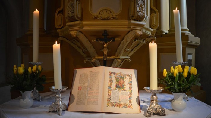 Ein Altar mit brennenden Kerzen und einem aufgeschlagenen Buch, umgeben von Blumenarrangements