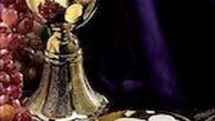 A golden chalice and paten are displayed with grapes and a purple cloth.