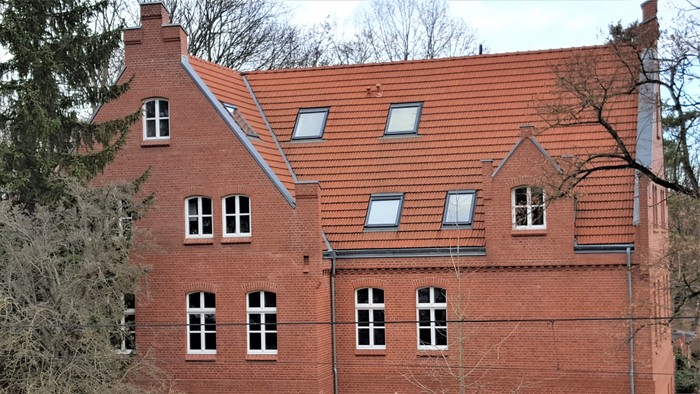 Das Bild zeigt ein zweistöckiges rotes Backsteingebäude mit mehreren Fenstern und einem roten Ziegeldach.