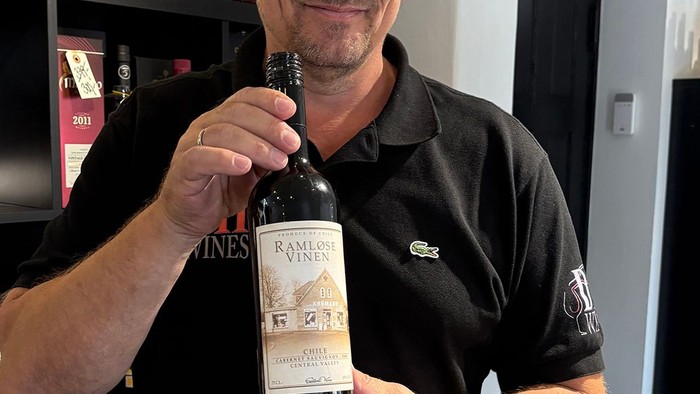 En mand i en butik holder en flaske vin op, omgivet af hylder med forskellige vine og spiritus.