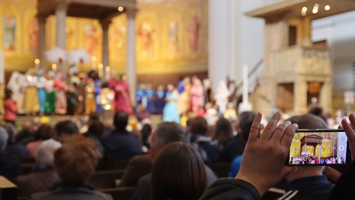 Eine Menschenmenge hält eine lebhafte religiöse Prozession in einer prächtigen Kirche fest, wobei Leute filmen und zuschauen.