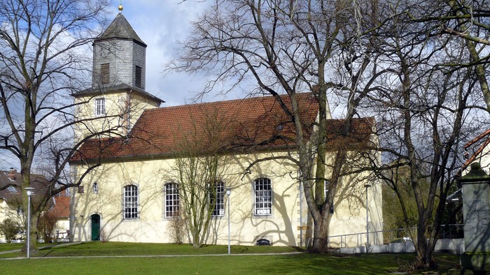 Alte Kirche mit rotem Dach und Turm, umgeben von kahlen Bäumen und grünem Gras