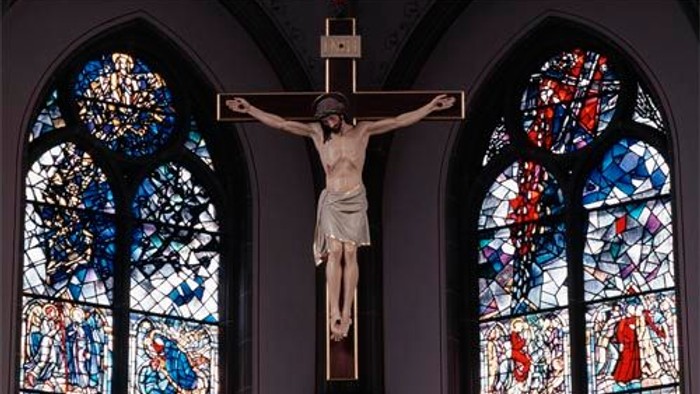 Das Bild zeigt ein Kruzifix vor zwei bunt gefärbten Kirchenfenstern.