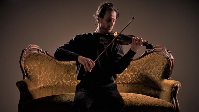 En mand spiller violin på en guldfarvet sofa.