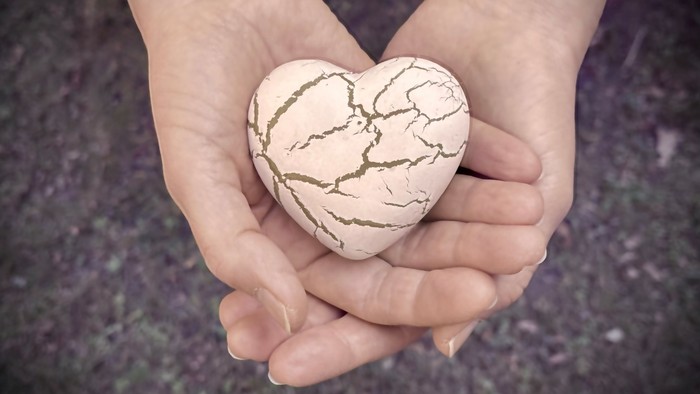 Hände halten sanft einen rissigen herzförmigen Stein, der Widerstandsfähigkeit und Liebe symbolisiert.