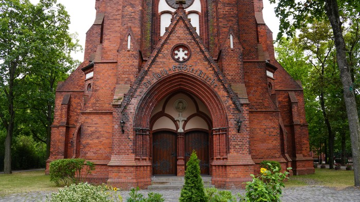 Eine historische Backsteinkirchenfassade mit einem bogigen Eingang, kunstvoller Steinmetzarbeit und üppigem Grün drumherum.