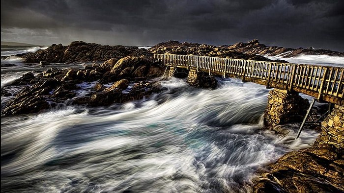 Klippekyst med bølger, der brænder mod klipperne, og en bro i baggrunden.