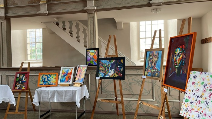 Kunstausstellung mit Gemälden auf Staffeleien in einem Raum mit Treppe.