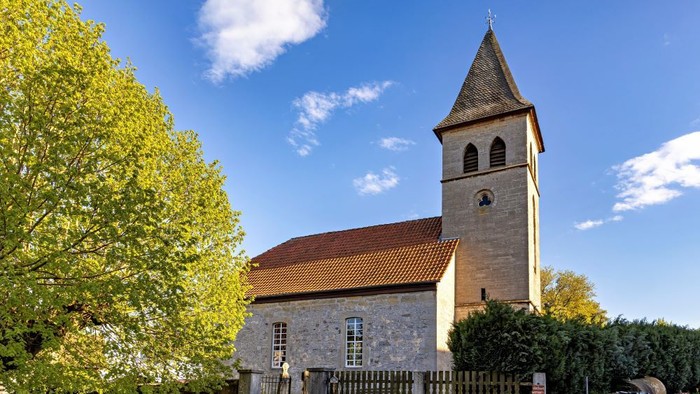 Kleine Kirche mit hohem Glockenturm, umgeben von Bäumen und einem Zaun.