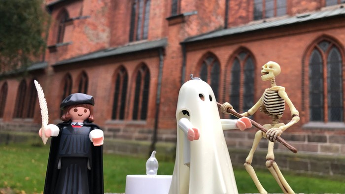Drei Spielzeugfiguren vor einer Kirche: ein Mönch mit Messer, ein Geist und ein Skelett.