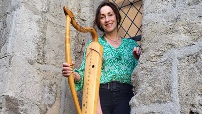 Eine Person hält eine große hölzerne Drehleier an eine Steinmauer gelehnt und lächelt.