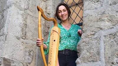 Eine Person hält eine große hölzerne Drehleier an eine Steinmauer gelehnt und lächelt.